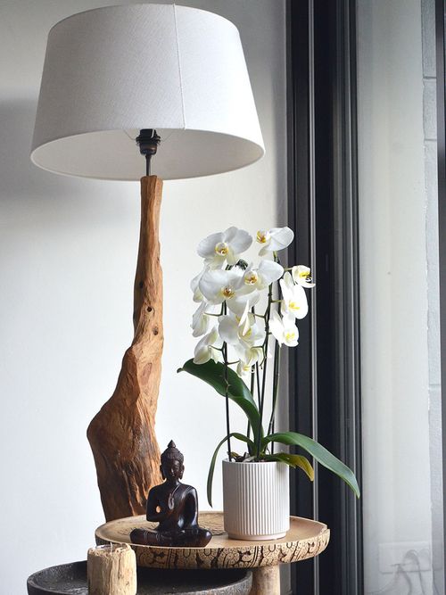 Ein kleiner Holztisch auf dem eine weiße Lampe mit einem Holzbein, ein Buddha und eine Orchidee steht.