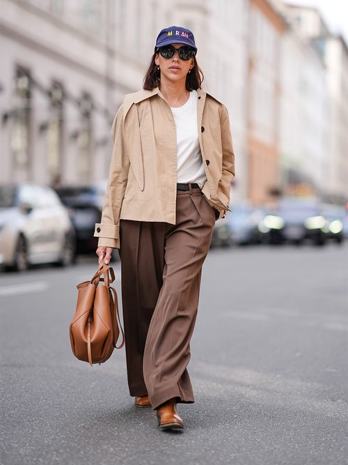 Street Style Look von der Copenhagen Fashion Week, wo ein Gast einen kurzen Trenchcoat, weißes T-Shirt und Bundfaltenhose trägt.