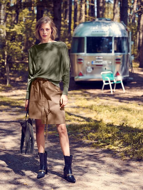 Frau im grünen Longsleeve zur beigen Rock steht vor einem silbernen Wohnwagen.