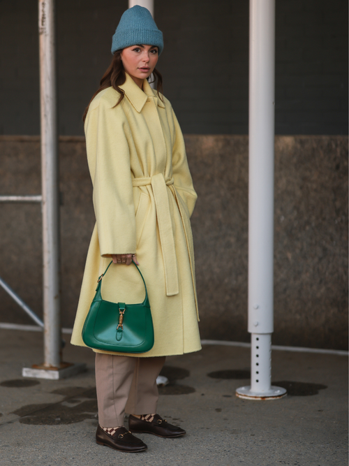 Street Style Look von der New York Fashion Week, wo ein Gast karierte Socken zu Loafers trägt. Mit einem gelben Mantel, grüner Tasche und beiger Hose.