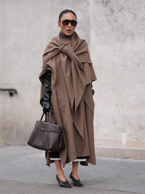A guest wears black sunglasses, dark brown draped knotted coat, shiny black leather gloves, dark brown leather bag, shiny black pointed toe heels, outside Ganni, during the Paris Fashion Week Spring/Summer 2025 on September 24, 2024 in Paris, France.