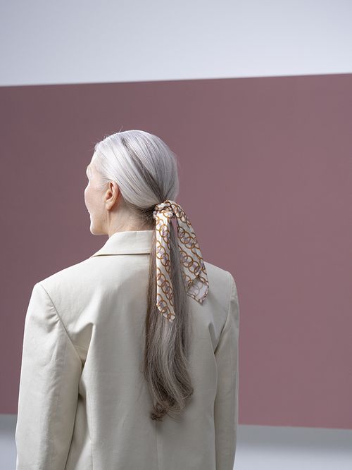 Frau mit einem beigen Blazer trägt einen Pferdeschwanz der mit einem Tuch gebunden ist.