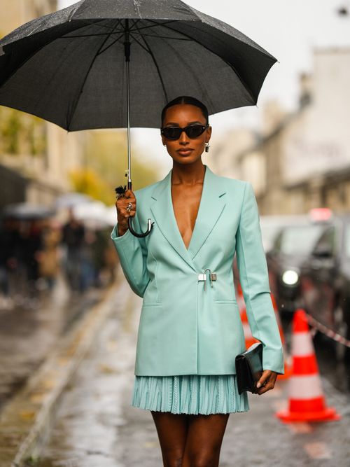 Street Style Look von der Paris Fashion Week, wo ein Gast ein türkies Ensemble aus Blazer und Faltenrock mit Regenschirm trägt.