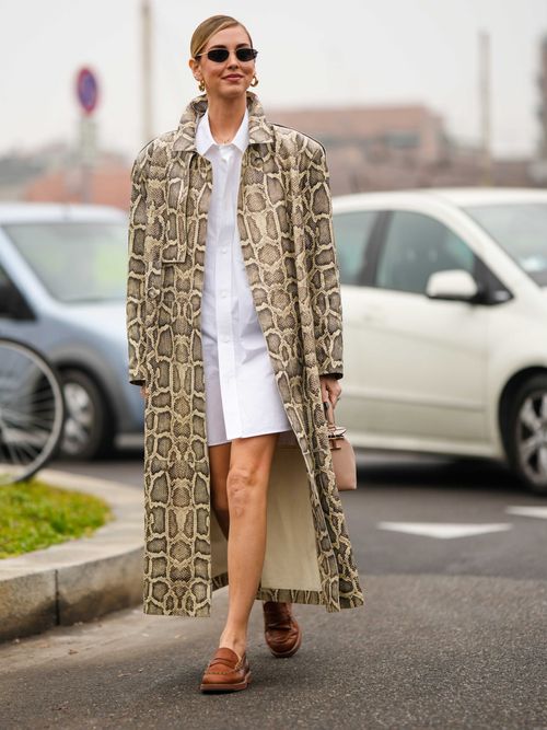 Chiara Ferragni wears black sunglasses, a white shirt short dress, a beige and brown shiny leather snake print pattern trench coat, camel leather loafers, a beige leather handbag, and gold earrings.