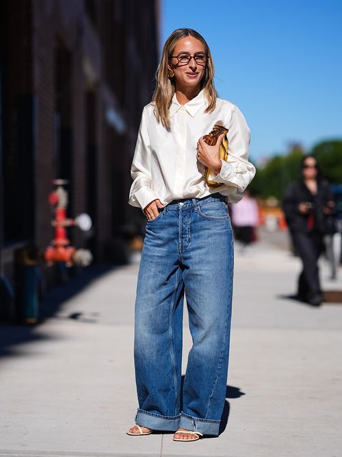 A guest wears black glasses, cream button up long sleeve shirt, shiny golf leather bag, washed navy blue denim jean cuffed pants, white strappy open toe heels, outside Ulla Johnson, during the New York Fashion week Spring/Summer 2025 on September 08, 2024 in New York, New York.