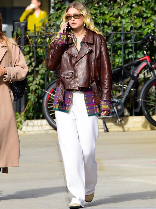 Gigi Hadid is seen in New York City wearing light brown sunglasses, dangling earrings, a brown leather jacket, a multicoloured tartan shirt, white wide-leg trousers, and cream suede shoes.