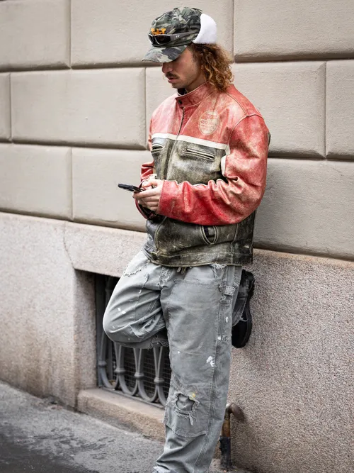 A guest wears red and olive green leather decorated jacket, grey distressed jeans, black shoes, military print hat, outside Giorgio Armani, during the Milan Fashion Week Menswear Fall/Winter 2025-2026 on January 20, 2025 in Milan, Italy.