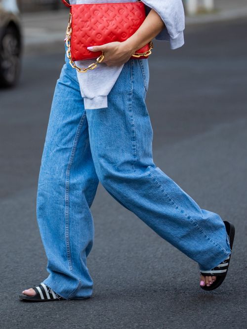 Sonia Lyson trägt einen grauen Pullover, eine weite Jeans, rote Tasche und Sonnenbrille mit Adiletten in Berlin.