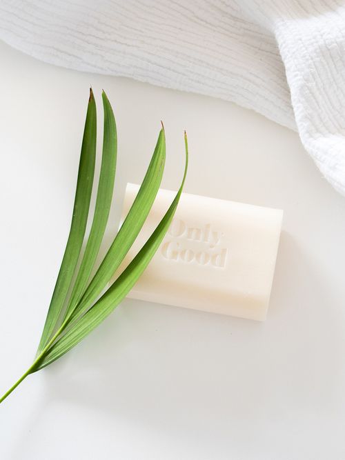 Ein Stück weiße Seife und ein weißes Handtuch liegen mit einem Blatt auf einer weißen Oberfläche.