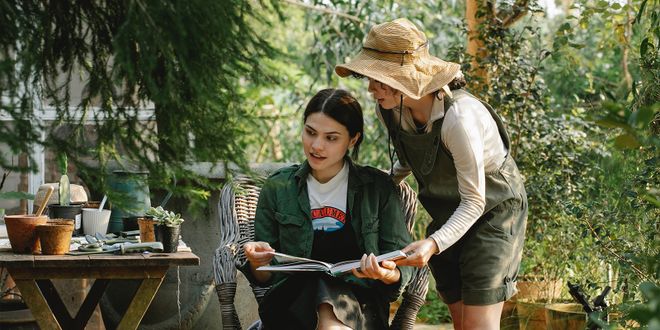 Zwei Frauen in khakifarbener Kleidung, sind im Garten und blättern in einem Buch.