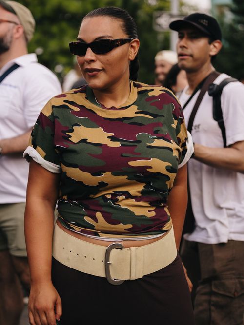 Paloma Elsesser is seen wearing black sunglasses, a camo print t-shirt, a wide cream suede belt, and black trousers.