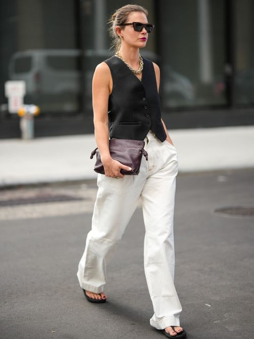 A guest wears black sunglasses, a large gold necklace, a black sleeveless / buttoned gilet, white latte denim large pants, a brown shiny leather clutch, black flip flop outside Tibi during New York Fashion Week.