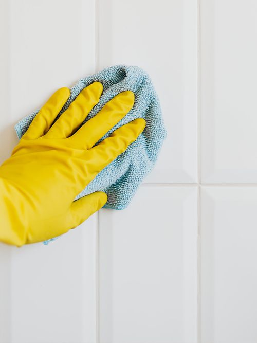 Hand mit einem gelben Gummihandschu und einem blauen Lappen, die über weiße Fließen wischt.