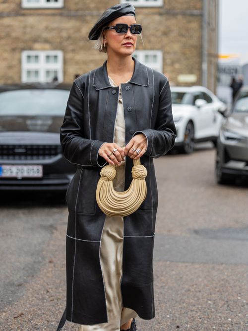 Street Style Look von der Copenhagen Fashion Week, wo ein Gast einen Leder-Trenchcoat mit Satinkleid und Barett trägt