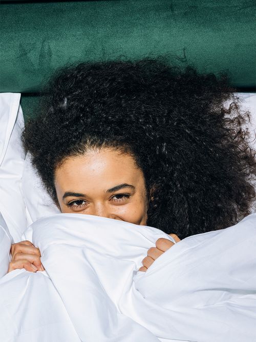 Frau mit lockigem schwarzen Haar liegt unter einer weißen Bettdecke, die sie sich halb über den Kopf zieht und lächelt.