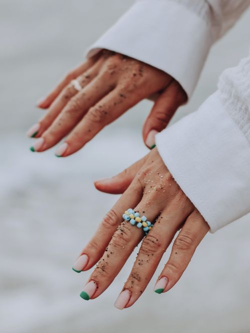 Two hands with green and nude abstract nail art and a yellow and blue beaded ring. The cuffs of a sheer white shirt are visible.