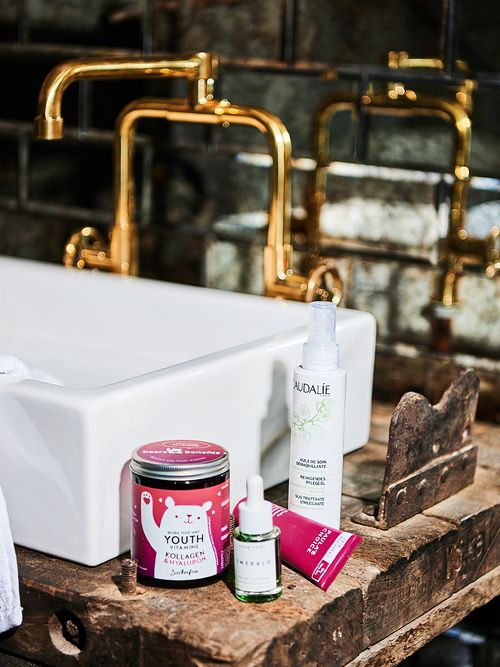 Weißes Waschbecken mit goldenem Wasserhahn vor dem Beautyprodukte stehen.