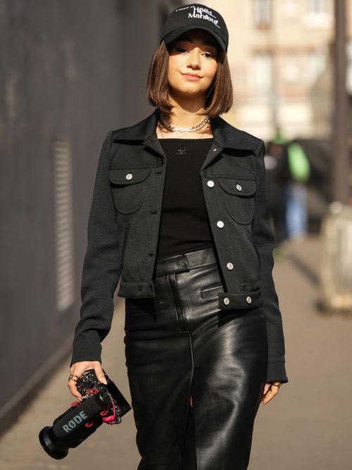 Lena Mahfouf trägt eine schwarze Jacke mit schwarzem T-Shirt und schwarzen Lederrock und Capy auf der Paris Fashion Week.