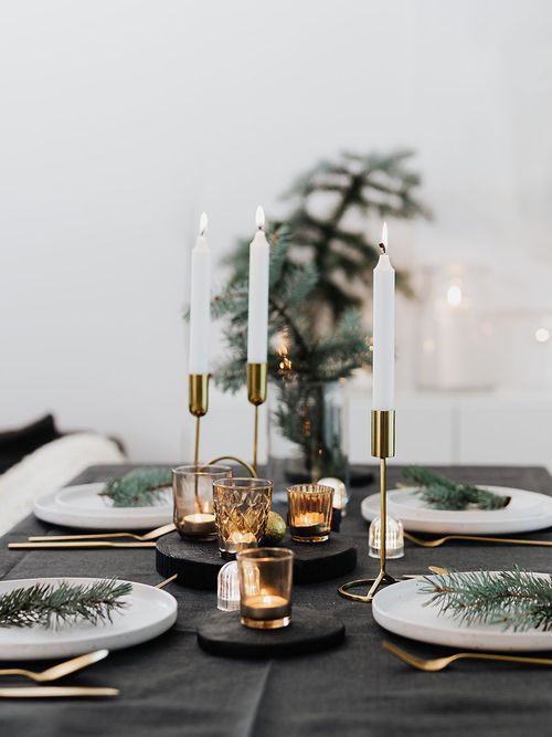 Ein weihnachtlich gedeckter schwarzer Tisch mit weißen Tellern, goldenen Kerzenständern und Tannenzweigen.