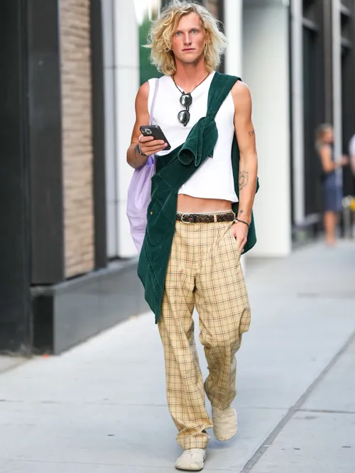 A NYFW guest wears a white tank-top, a dark green velvet shirt, a pale purple shoulder bag, black sunglasses, silver rings, a watch, a black and brown braided shiny leather belt, pale yellow with black and red checkered print pattern large pants, and beige suede sneakers.