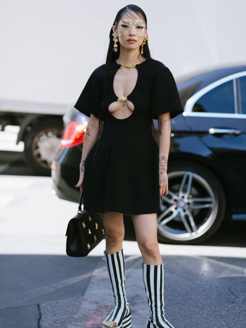 Rina Sawayama wears a black cut-out mini dress, an embellished black leather bag, and black and white knee-high boots with golden toe details at Schiaparelli's FW23 show during PFW.