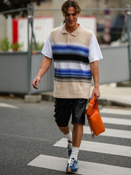 A guest wears a beige with black and blue striped print pattern with white short sleeves oversized polo shirt, black shiny leather shorts, an orange shiny leather handbag, beige socks, blue denim ripped sneakers.