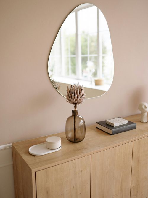 Brown side table with a white cup, a brown vase, a blue book, and a mirror hanging on the wall.
