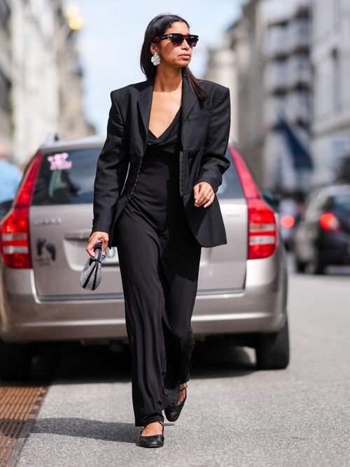 Miriam Akosua Nehring wears black sunglasses, white earrings, black tailored blazer jacket, black maxi dress, silver sequin black bag, gold rings, shiny black ballerina flat leather shoes, outside Gestuz, during the Copenhagen Fashion Week Spring/Summer 2024-2025 on August 8, 2024 in Copenhagen, Denmark.