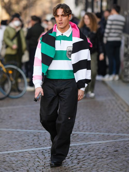 A guest wears a green and white striped print pattern long sleeves polo shirt, a pink and black striped print pattern pullover, black large pants, black shiny leather shoes , outside MSGM, during the Milan Menswear Fall/Winter 2023/2024.
