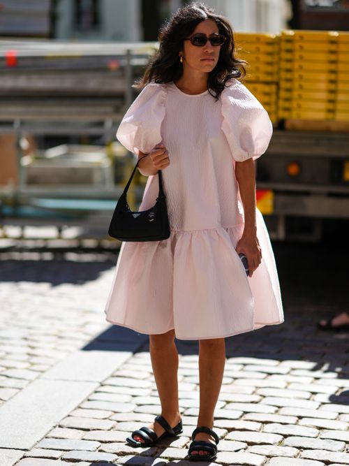 A guest wears black sunglasses, gold earrings, a white puffy short sleeves / ruffled knees dress, a black nylon handbag from Prada, black shiny leather strappy sandals.