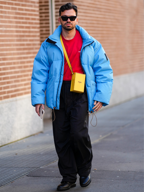Ein Milan Fashion Week Gast trägt eine blaue Daunenjacke mit einem rotem T-Shirt, schwarzer Hose und gelber Tasche