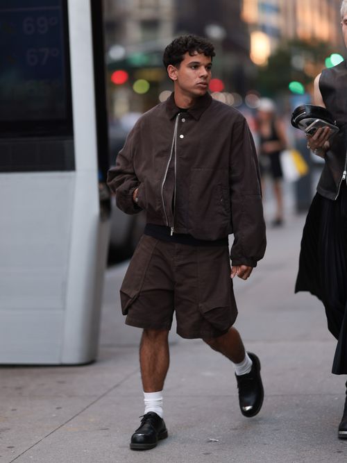 A Fashion Week guest is seen wearing two tiny stud earrings; a dark brown blouson jacket with a corduroy collar, puffy sleeves, a silver zipper, and two silver buttons from Jacquemus; a black T-shirt underneath; knee-length dark brown cargo shorts with large pockets along the sides from Jacquemus; white socks; and black leather three-eye shoes with an exposed silver steel toe cap from Dr. Martens before the Jacquemus Boutique Store Opening during the New York Fashion Week on September 04, 2024 in New York City.