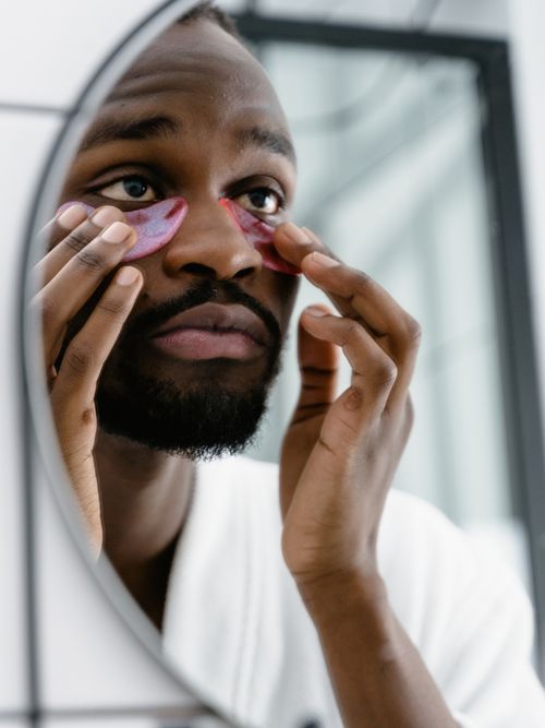 Mann mit Bart steht vor dem Spiegel im Badezimmer und trägt Eyepatches auf