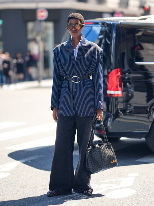 Street Style Look von der Paris Fashion Week, wo ein Gast einen gestreiften Blazer mit Taillengürtel, ausgestellte Hose, Sonnenbrille und Handtasche trägt.