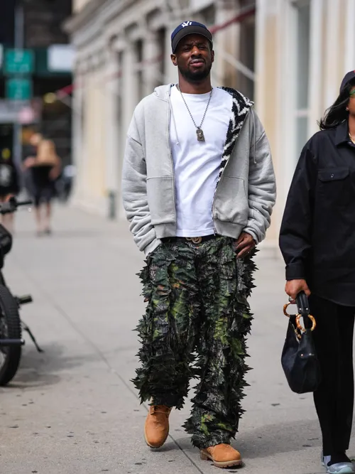 A guest wears navy blue New York Yankees baseball hat, white t-shirt, silver necklace, light gray oversized unzipped hoodie, black belt, dark gray green camouflage graphic detail pants, light brown boots, outside Who Decides War, during the New York Fashion week Spring/Summer 2025 on September 07, 2024 in New York, New York.