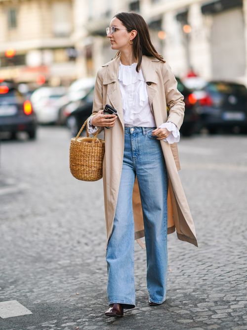 Marielle Haon trägt einen beigen Trenchcoat mit weißer Bluse und Jeans, sowie einer Korbtasche.