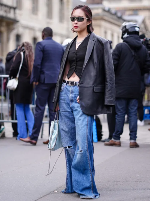 A guest wears black sunglasses, a black buttoned sleeveless gilet, a black shiny leather blazer jacket, a black shiny leather belt, blue denim faded ripped wide legs pants, a pale blue shiny faux leather handbag from Stella McCartney.