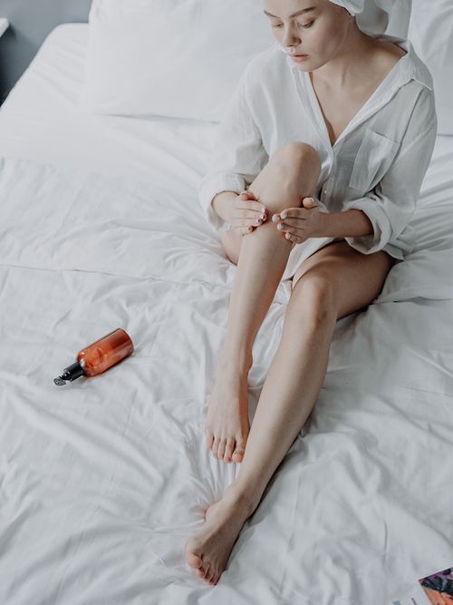 Frau in weißem Männerhemd cremt sich am Bett die Beine ein.