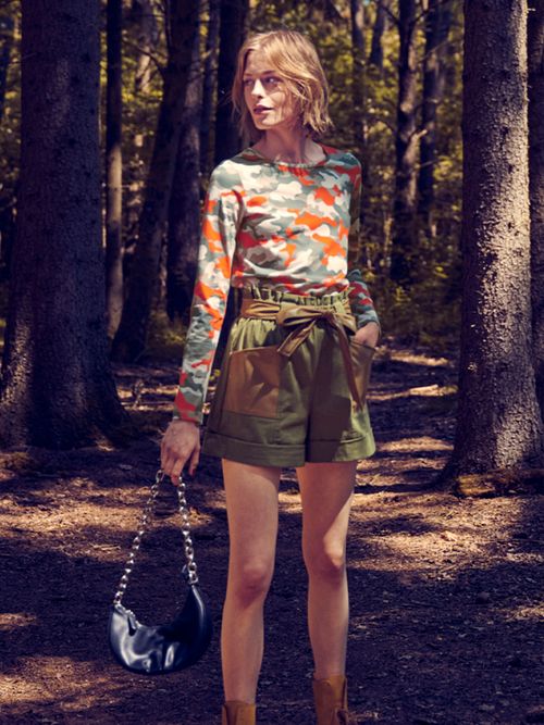 Frau steht im Wald mit einer khakifarbenen Shorts und einem Longsleeve mit Camouflage-Print.