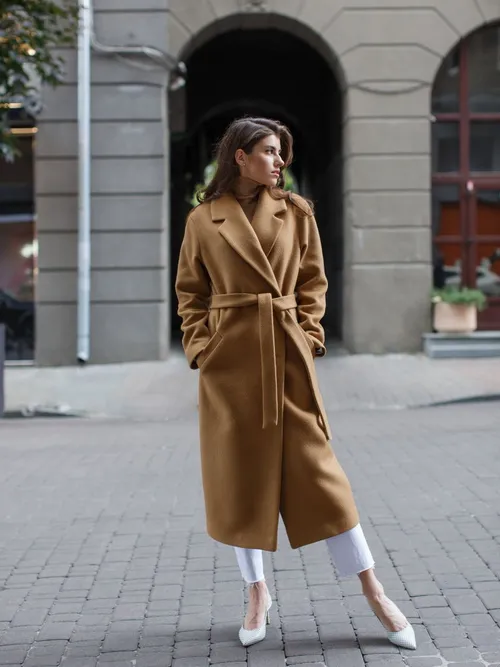 Street Style Look von einer Frau, die einen beigen Wollmantel mit Gürtel, weißer Hose und weißen High Heels trägt.