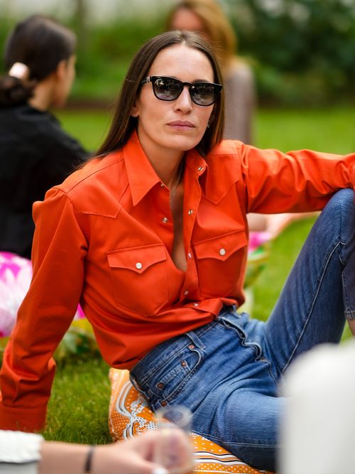 Frau in Jeans und einer orangefarbenen Bluse sitzt im Gras.