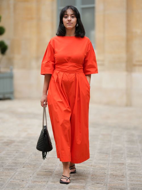 Street Style Look aus Paris, wo eine Frau einen orange-rotfarbenen Jumpsuit mit schwarzer Tasche und Sandalen trägt.