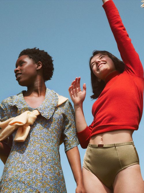 Zwei Frauen die lachend vor einem blauen Himmel stehen. Eine mit rotem Pulli und eine in einem grauen Oberteil.