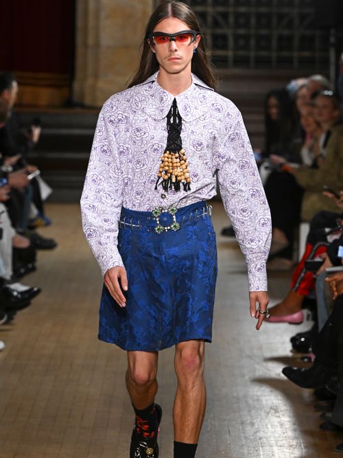 A Chopova Lowena model wears orange sunglasses, a printed button-up shirt, a metal bolo tie, a denim knee-lenght skirt, black and red socks, and black loafers.