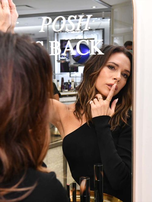 Victoria Beckham in einem schwarzen Kleid steht vor einem Spiegel und hält sich den Zeigefinger vor die Lippen.