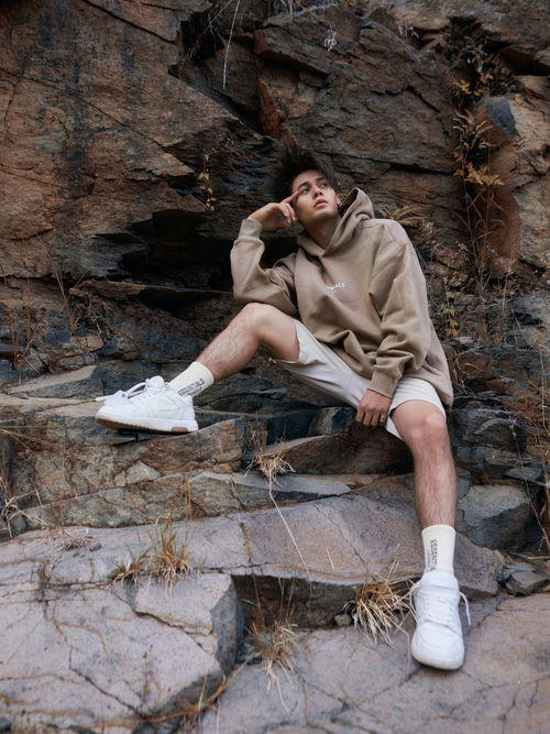 A man wearing a beige hoodie, khaki shorts, and white sneakers sits on a rock.