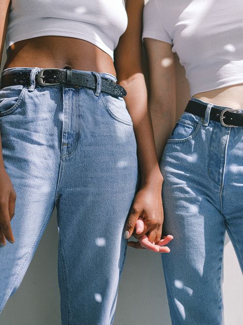 Zwei Frauen in hellen Jeans mit schwarzen Gürtel und weißem Top, die Händchen halten.