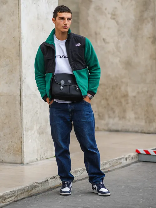 Street Style Look von der Paris Fashion Week, wo ein Gast eine North Face Jacke in Grün, ein weißes T-Shirt, Jeans, Nike Sneaker trägt und Umhängetasche trägt.