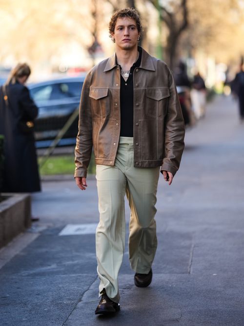 Street Style Look von der Milan Fashion Week, wo ein Mann eine Lederjacke, schwarzes Poloshirt und beige weite Hose trägt.