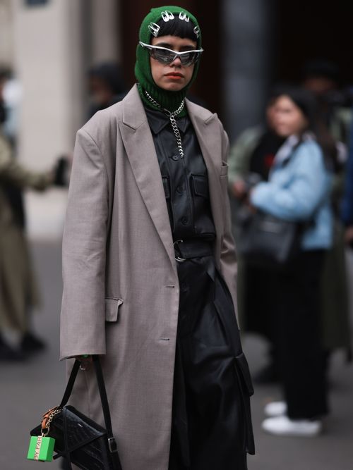 A guest wears a green balaclava, silver metallic hair clips, silver sunglasses, a black leather dress, a taupe blazer coat, a black leather bag and a silver chain, outside before Palm Angels Show, during Paris Fashion Week on March 05, 2023 in Paris, France.